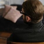 A man with glasses sits contemplatively indoors, suggesting a reflective or therapeutic moment.